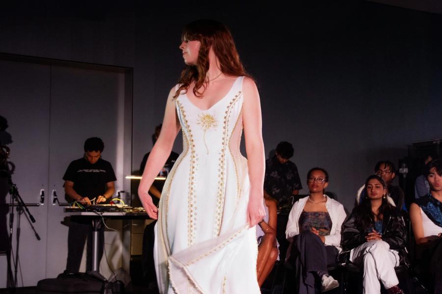 Model walks in a white dress featuring gold embroidery and a sunburst motif on the bodice. The structured gown has a sweeping hemline and is framed by seated spectators.